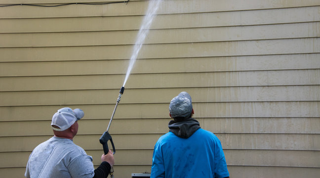 Two Young Men Standing By The Side Of A Yellow House. One Is Power Washing The Siding With A Long Nozzle.