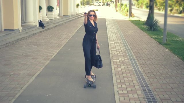 Modern business woman in a black jumpsuit in beige shoes , sunglasses and a black bag riding on an electric board along a city street, depicting a super man, stretching one arm forward. The concept of