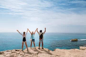 Gordijnen Het gezelschap van vrienden van reizigers staat aan de rand van een klif tegen de achtergrond van de zee. Ze staken hun handen omhoog. Cyprus. © kazantsevaov