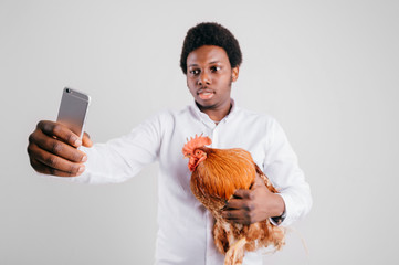 Young funny african american man with rooster in hands taking selfie on white background.