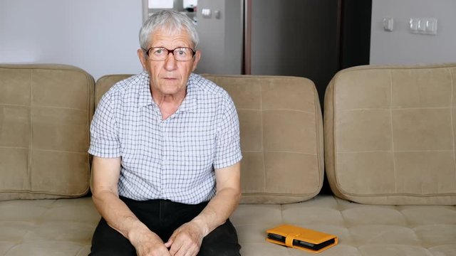 Elderly European puts on glasses sitting on a sofa. An elderly man is alone in a room, sitting on a couch.