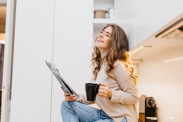 Laughing girl in beige shirt reading news while drinking coffee in morning. Indoor portrait of...