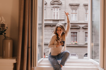 Joyful smiling woman sitting on sill and waving hand. indoor portrait of fashionable young lady with curly hair posing near window with cup of hot bevegare.