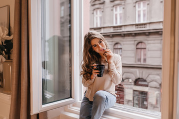 Romantic dark-haired girl spending time at home and drinking latte on sill. Portrait of dreamy caucasian young woman wears shirt and jeans posing next to window.
