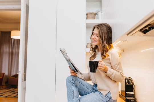 Joyful dark-haired lady spending morning at home, reading newspaper with smile. Indoor photo of carefree girl in jeans enjoying tea in kitchen.