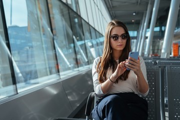 Young passenger woman in casual clothes sitting at the gate and using smartphone while waiting for a flight at the airport. Travel concept. Girl chatting in terminal departure lounge.