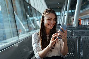 Traveler woman sitting at the gate and using smartphone while waiting for a flight at the airport. Travel concept. Young passenger girl chatting and smiling in terminal departure lounge.
