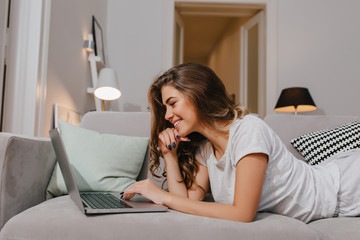 Amazing curly girl with black manicure lying on couch and typing on laptop keyboard. Smiling young...