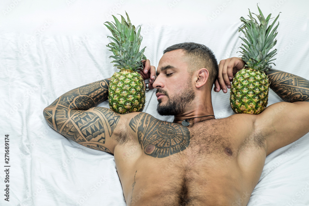 Wall mural muscular man hold pineapple fruit on biceps. brutal sportsman relax in bed. steroids. sexy ab of tat