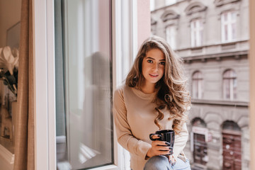 Fototapeta na wymiar Interested shy girl with long hair posing with cup of tea on sill. Indoor portrait of good-looking curly model in beige shirt sitting near window on city background.