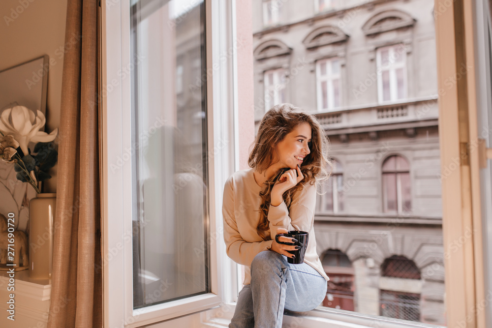 Wall mural romantic female model in good mood looking at street, sitting on sill. fascinating dark-haired lady 