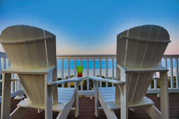 relax on Adirondack chairs in a desk balcony overlooking the beach and  ocean at sunset.  Add a beverage to this travel image.