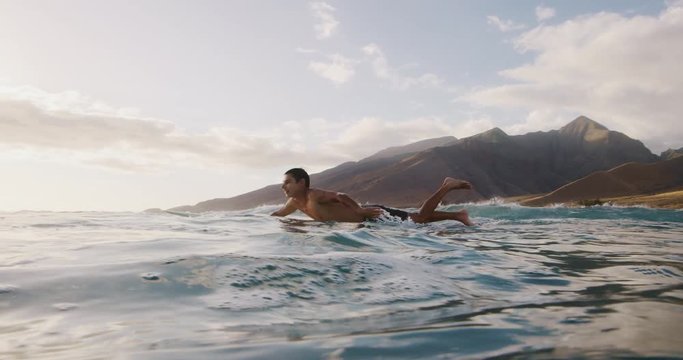 Young man surfing at sunset, paddling out on his longboard with island in the background