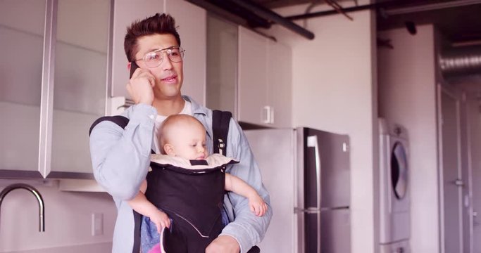 Young millennial stay at home dad working on phone while holding baby daughter 
