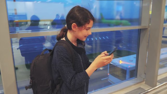 daughter with a backpack airplane flights airport concept. girl teenager with a smartphone looks out the window at the airport area airplanes and bus at a distance. teen girl looks at lifestyle the