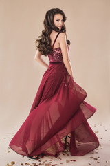 Gorgeous brunette curly hair model posing in elegant burgundy evening dress on the beige paper...