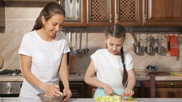 Portrait of mother and daughter are cooking apple pie together in the kitchen, mom is kneading a dough and her teen daughter is cutting apple.