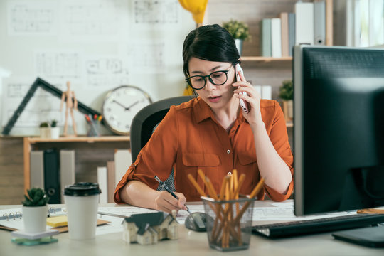 young asian female engineer worker sitting at desk in studio talking on cellphone with boss writing down note on paper. elegant lady architect in glasses listening concentrated focus on smart phone