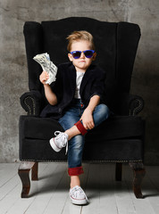 Young rich boy kid millionaire sits in big luxury armchair and demonstrates bundle of money dollar bills