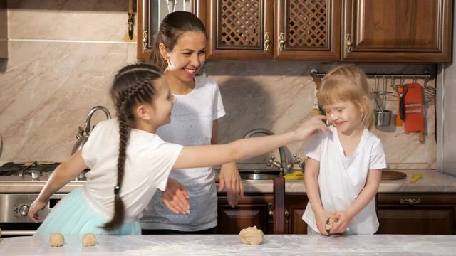 Portrait of playful mother and daughters are cooking dough for pie together in the kitchen and smearing each other in flour, touching noses with dirty hands, funny time together.