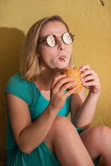 A happy woman in a turquoise dress sits on the sidewalk and eats a burger on a warm summer day. Beautiful blonde in sunglasses enjoys junk food at the yellow wall of a residential building.