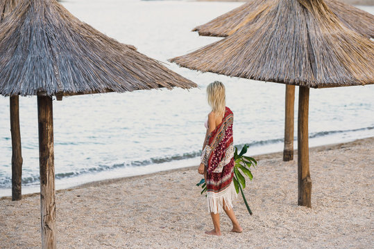 woman in boho style on the beach stands near the water and holds exotic leaf