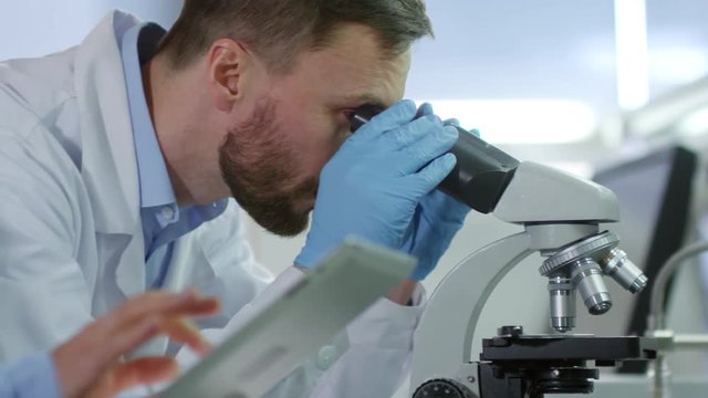 Close up of male biochemist using microscope and telling investigation results to his female laboratory assistant writing them down using tablet