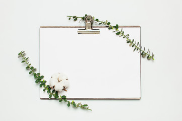 Fototapeta na wymiar Blank white paper with folder and metal clothespin, spring green twigs of plants on gray background top view flat lay copy space. Decorative plant branch, flowers composition mockup Minimalistic style
