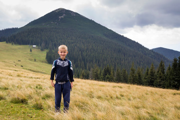 Fototapeta na wymiar Young happy smiling child boy with backpack standing in mountain grassy valley on background of summer landscape, woody mountain view. Active lifestyle, adventure and weekend activity concept.