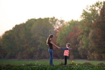 Fototapeta premium Back view of young slim attractive mother and child girl standing in green meadow holding hands outdoors on forest trees blurred background.