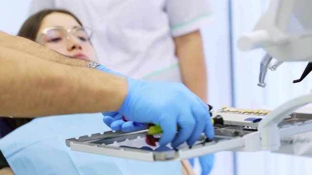 Young woman is waiting in dentist chair while dentist is preparing tools for filling her tooth, putting them on metal tray and choosing the color of the seal from samples.