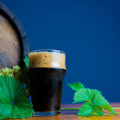 glass of stout on a wooden table with leaves of hop against the background of barrels