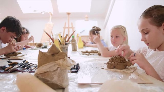 Children are sculpting toys from wet clay. They are using special tools under the guidance of a teacher on art lesson in workshop in elementary school.