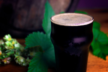 Pint of dark beer foam beautifully on a barrel with a leaf of hops background