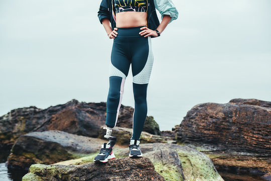 Cropped photo of disabled woman in sports clothing with prosthetic leg standing on the stone and keeping arms on her hips.