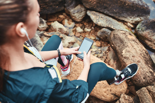 Disabled woman in sport wear with leg prosthesis holding phone in her hand and listening music while sitting on the boulders - Powered by Adobe