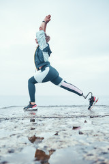 Surya Namaskar. Vertical photo of disabled athlete woman in sportswear with prosthetic leg standing in yoga pose on the stone in front of the sea.