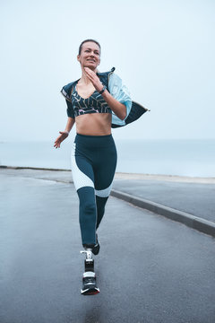 Can't live without sport. Vertical photo of strong disabled woman in sportswear is running outdoors near the sea and smiling.
