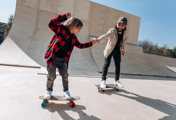 Father and his little son dressed in the casual clothes ride skateboards in a skate park with...