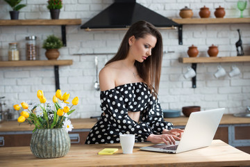Attractive brunette girl blogger in a polka dot dress sitting in the cozy kitchen with laptop at the wooden table