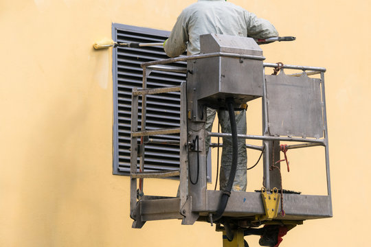 Man On A Lifting Platform Painting The Building Wall With A Roller Exterior Outdoors. Worker On A Ladder Manually Painting Yellow Wall On Construction Site