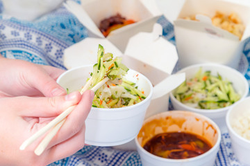 Eating Chinese take away food with chop sticks at home with bed sheets in the background. Spicy asian food in white box - salad, souse, rice with egg, dry beef, crunchy chicken cooked in oil