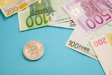 Silver Bitcoin coin next to Euro bank notes on blue background. Digital currency, block chain market. Euro bills next to crypto coin