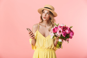 Beautiful thinking amazing young blonde woman posing isolated over pink wall background holding flowers using mobile phone.