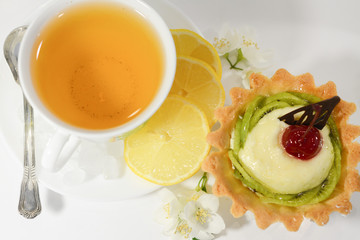 A mug of green tea with lemon and sugar and a cream dessert with kiwi and cherry on top in a basket of shortcake pastry on a white background