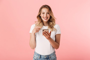 Happy young blonde woman posing isolated over pink wall background using mobile phone.
