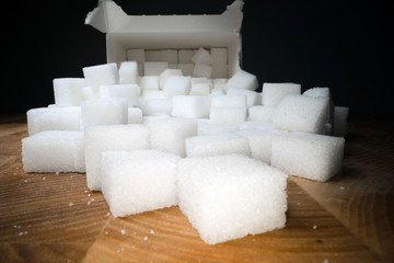 Macro shot of sugar cubes next to carton box on wooden table. Unhealthy food sweetener, sweet crystal cubes