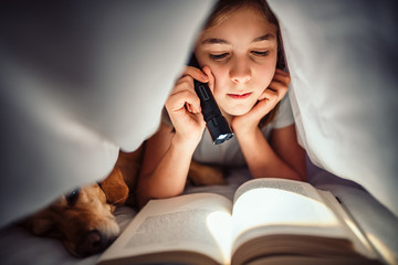 Girl lying in the bed with her dog under blanket reading book late at night