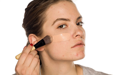 Young woman with problematic skin applying liquid foundation on white background