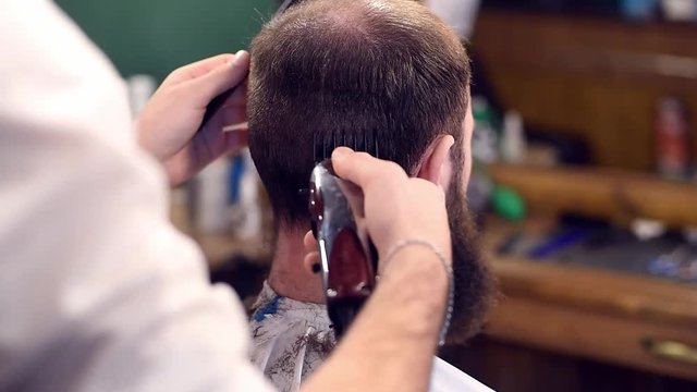 Close up filming of making male haircut with electric razor. Cropped barber's hands combing out and cutting short hair on back of head for brunette bearded invisible male client. Blurred background